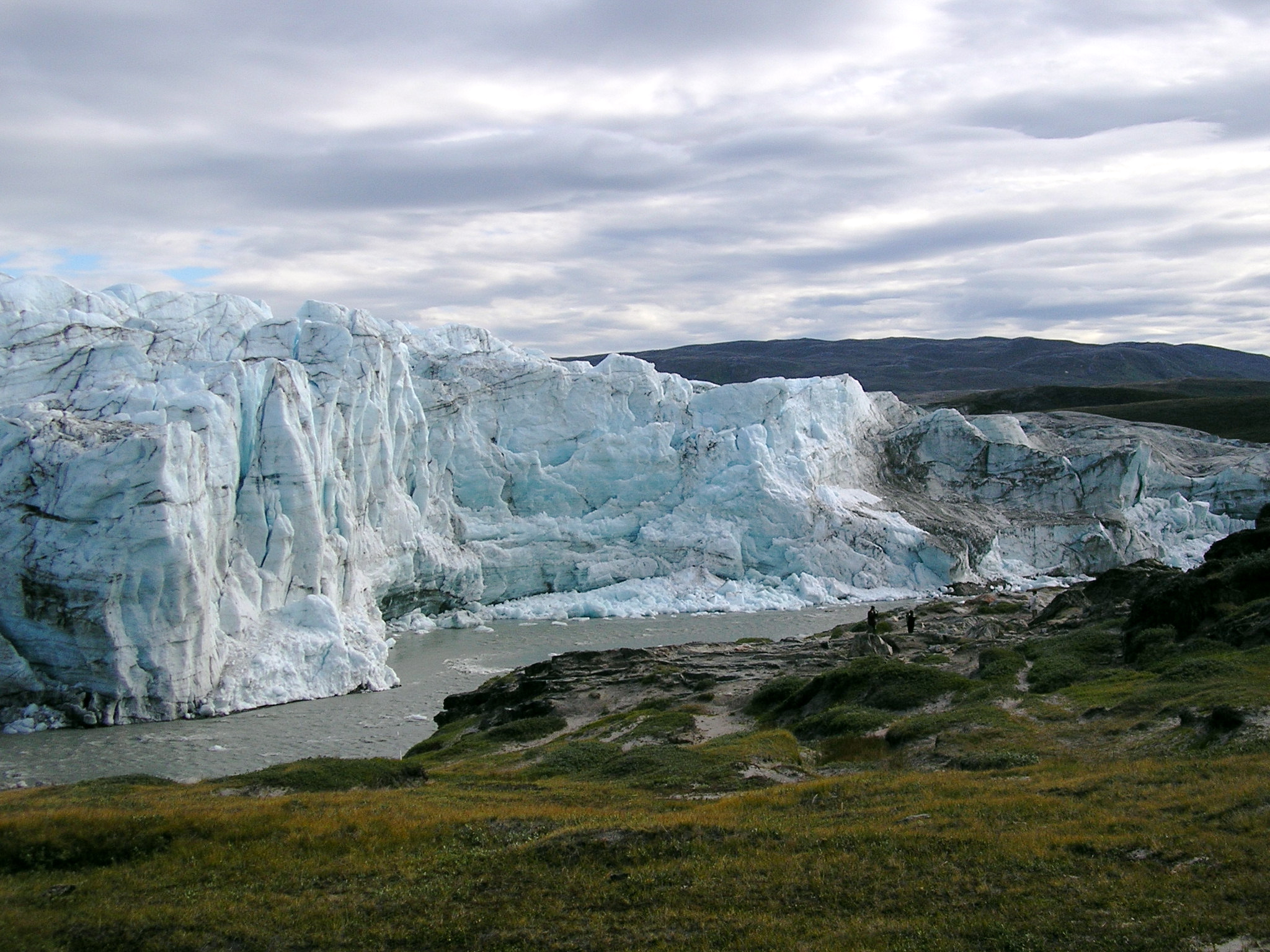 Photo via WikiMedia Commons, The edge of the Greenland icesheet, note the humans standing at the center right hand side for scale. Taken near Kangerlussuaq, Greenland; 8-11-2005 by L. Chang